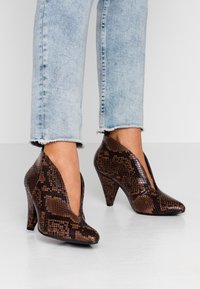 Brown snake-patterned high-heeled shoes with a pointed toe and side cutouts, paired with light blue frayed hem jeans.
