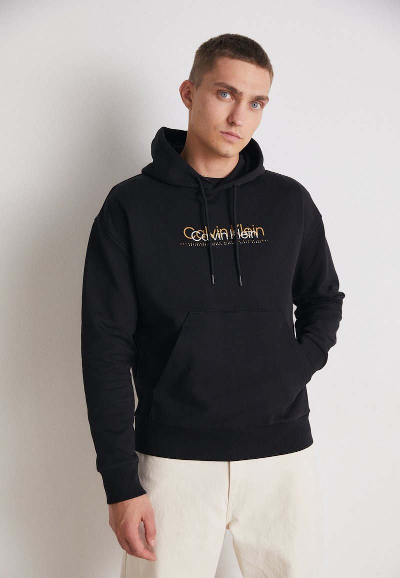 Felpa nera in cotone con tasca frontale, cordino regolabile e logo Calvin Klein dorato. Texture liscia e polsini aderenti.