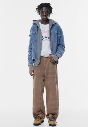 Das Model trägt eine blaue Cordjacke mit grauer Kapuze, ein weißes T-Shirt mit Grafik, braune weit geschnittene Hosen und gelbe Stiefel mit blauen Schnürsenkeln.