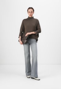 Poncho en tricot marron avec un col haut, associé à un jean bleu clair évasé. Le mannequin tient un sac à main structuré marron foncé.