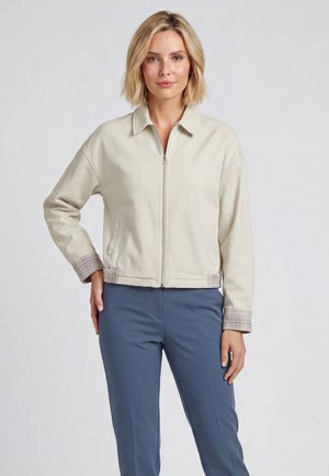 Frau mit kurzen blonden Haaren, die eine beige Jacke mit Reißverschluss und karierte Bündchen sowie blaue Hosen trägt, stehend vor einem schlichten hellen Hintergrund.