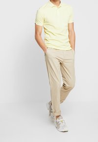Polo giallo con maniche corte, abbinato a pantaloni chino beige e sneakers grigie. Dettagli notevoli includono un sottile logo del marchio sulla maglietta.