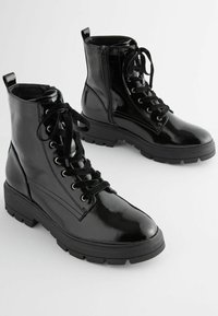 Bottines en vernis noir à bout rond, avec lacets à l'avant, fermeture éclair sur le côté et semelle robuste. Présentent une texture lisse et un design solide.
