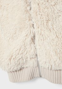 Chaqueta esponjosa color crema con un zipper plateado parcialmente desabrochado, que muestra la cintura de punto acanalado en la parte inferior.