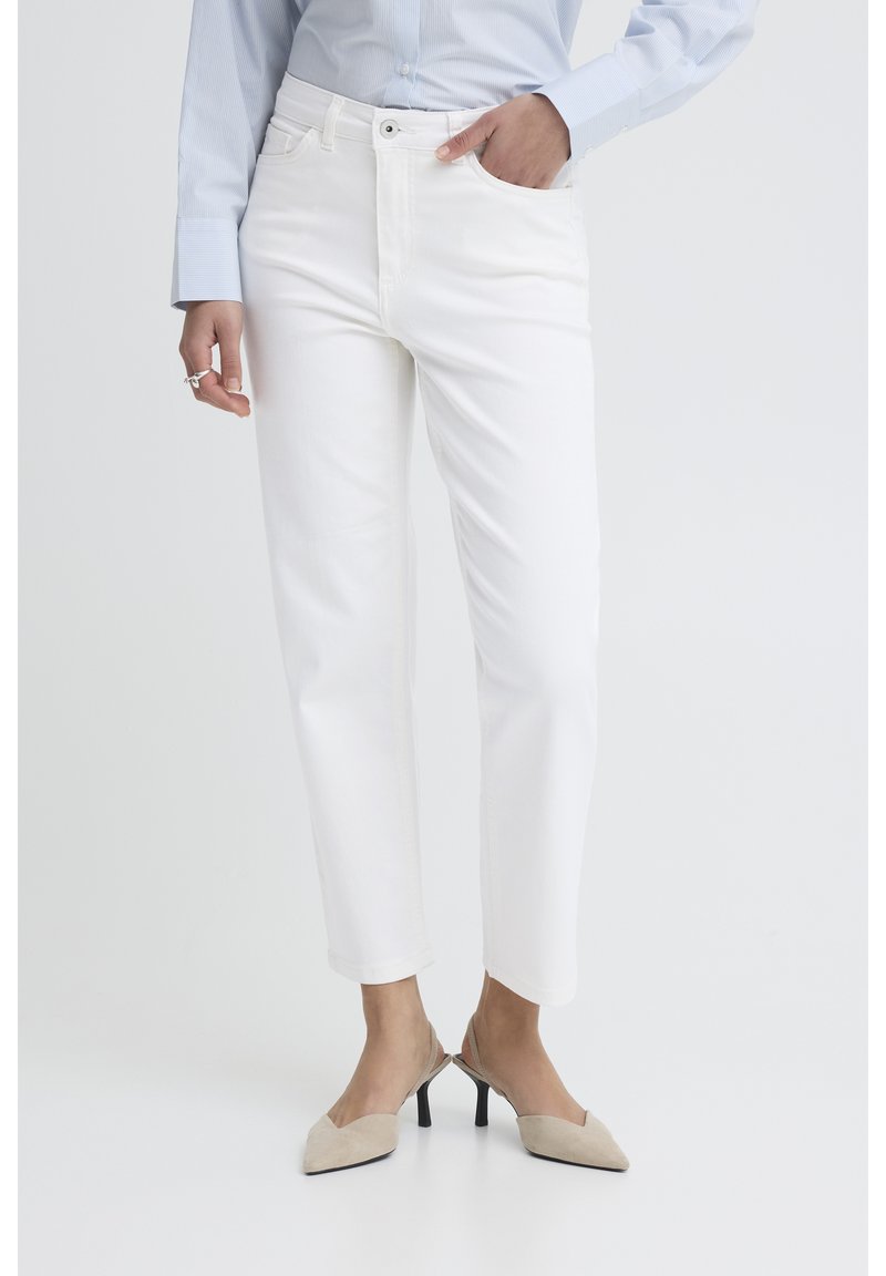 Femme portant un pantalon blanc à jambes droites, des talons slingback beige, une chemise bleu clair à manches longues, avec une main dans la poche et des bagues aux doigts.