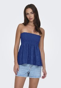 Brezramni modri top z teksturirano, nabrano tkanino in eyelet detajli, v kombinaciji z svetlimi jeans kratkimi hlačami s svetlobnim vzorcem zvezd.