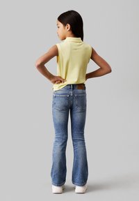 Žluté bezrukávové polo tričko s límečkem, kombinované s modrými rozšířenými džínami. Džíny mají zadní kapsy a koženou nášivku v pase.