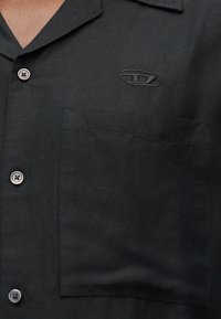 Κοντινό πλάνο μαύρου πουκαμίσου με ασημένια κουμπιά, τσέπη στο στήθος και μικρό κεντημένο οβάλ λογότυπο στην επάνω αριστερή πλευρά.