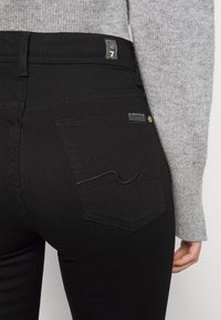 Jeans negros con una textura suave y ajuste slim, con bolsillos traseros que tienen detalles de costura y una pequeña etiqueta de logotipo de la marca.