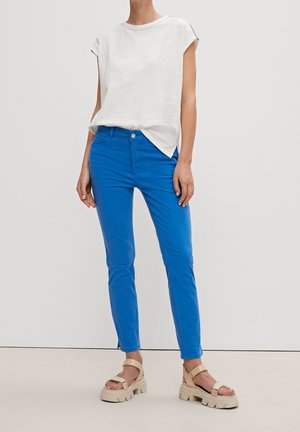 Kobieta ubrana w biały bezrękawnik, jasnoniebieskie dopasowane spodnie i beżowe masywne sandały na jednolitym jasnym tle.