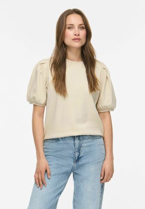 Donna con capelli lunghi e castani che indossa una maglia crema con maniche a sbuffo e jeans a vita alta azzurri chiaro in piedi davanti a uno sfondo bianco semplice.