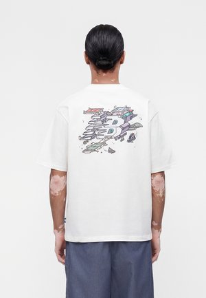 Osoba z włosami związanymi do tyłu nosząca białą koszulkę z kolorowym, abstrakcyjnym, warstwowym wzorem statku kosmicznego na plecach, w połączeniu z niebieskimi spodniami.