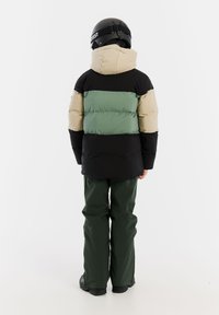Gepolsterte Jacke in Grün, Beige und Schwarz; ausgestattet mit einer Kapuze und elastischen Bündchen. Kombiniert mit grünen Hosen und schwarzen Skischuhen.