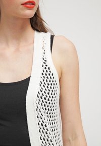 Gestrickte weiße Weste mit offenem Maschenmuster, getragen über einem schwarzen Tanktop. Glatte Textur und schichtiger Look, der die Haut darunter zeigt.
