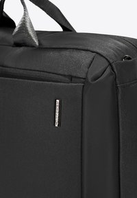 Sac en tissu noir avec une texture lisse et une finition brillante, fermeture éclair, et un accent de logo argenté sur le côté. Design compact.