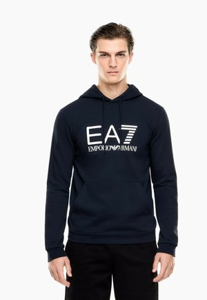 Ung mand iført en navy blå EA7 Emporio Armani hættetrøje med frontlomme og sorte bukser, stående mod en hvid baggrund.