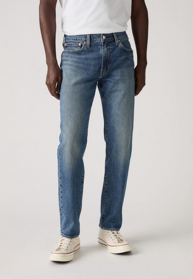 Homme portant un jean bleu délavé, des baskets blanches et un t-shirt blanc, debout devant un fond uni.