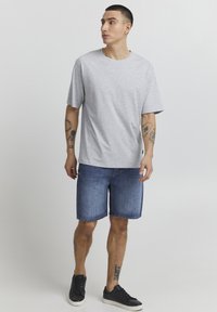 T-shirt gris à manches courtes, coupe décontractée, associé à un short en denim bleu et des baskets noires. Tatouages visibles sur les bras, fond uni.