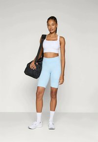 Ανοιχτό μπλε σορτσάκια ποδηλασίας και λευκό crop top, με μια μαύρη τσάντα γυμναστηρίου κρεμασμένη στον έναν ώμο και λευκά αθλητικά παπούτσια. Λείες υφή, εφαρμοστός σχεδιασμός.