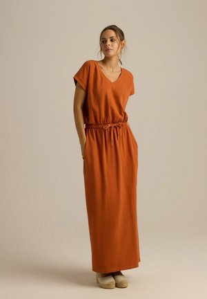 Pomarańczowa, długa sukienka z krótkimi rękawami, dekoltem w serek, elastycznym pasem i kieszeniami. Materiał wydaje się być miękki; stylizowana z beżowymi espadrylami.