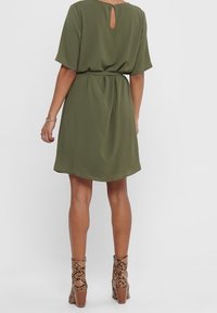 Oliivinvihreä, polvimittainen mekko, jossa on lyhyet hihat, avainreikätakaosa ja vyötärölle korostava muotoilu. Yhdistetty käärmeennahkakuosisilla korokepohjaisilla nilkkabootseilla.