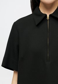 Top de tecido preto com um colarinho curto, mangas curtas e um fecho dourado no decote, apresentando uma superfície texturizada.