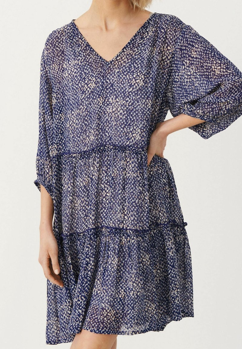 Femme portant une robe ample à motifs bleu et beige avec un col en V et des manches trois-quarts, tenant une main sur la hanche.