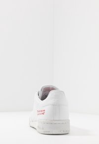 Sneaker in pelle bianca con punta arrotondata, suola piatta e dettagli testurizzati. Presenta testo rosso sul tallone e logo Adidas sul retro.