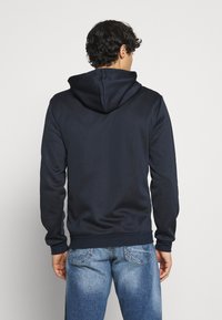 Marineblå hættetrøje med justerbar snor i hætten, blød tekstur og ribstrikkede ærmegab; kombineret med lyseblå denimjeans. Set bagfra.