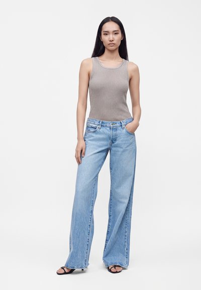 Femme aux longs cheveux foncés portant un haut gris sans manches, un jean bleu clair à jambes larges et des talons ouverts noirs, debout avec une main dans la poche.