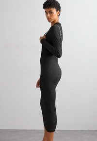 Abito nero in maglia a costine con maniche lunghe, silhouette aderente e orlo scallop. Accento notevole di catena sulla spalla.