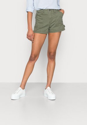 Femme portant un short vert olive avec des poches avant, une chemise bleu clair aux manches retroussées et des baskets blanches à plateforme, debout avec une main dans la poche.