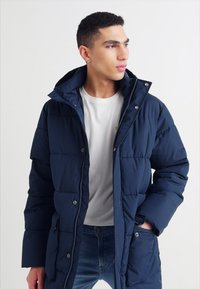Námořnická puffer bunda se zipem a patentkami, má kapuci, prošívaný design a boční kapsy. Nositá přes bílé tričko.