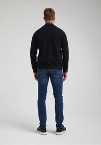 Felpa blu navy con texture e colletto, indossata sopra una maglietta grigia a maniche lunghe; abbinata a jeans slim-fit blu e sneakers nere.