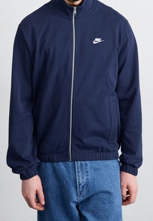 Mies pukeutuneena tummansiniseen Nike-vetoketjutakkiin, jossa on joustavat hihansuut, ja vaaleansinisiin farkkuihin, seisoo kädet rentoina sivuilla.