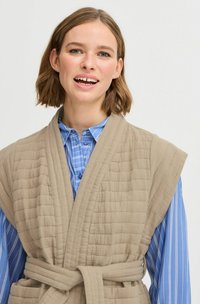Gilet en beige matelassé avec des manches larges, surface texturée et ceinture nouée, superposé sur une chemise rayée bleu et blanc.