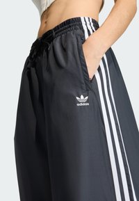 Pantalones deportivos negros con rayas blancas en los costados, cintura elástica y bolsillos. Hechos de tela ligera y suave. El logo de Adidas es visible.
