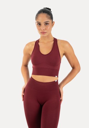 Femme athlétique aux cheveux foncés en chignon, portant un soutien-gorge de sport bordeaux sans coutures assorti et un legging taille haute, debout les mains sur les hanches.