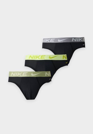 Мускулест мъж, изправен бос, носещ черни слипове на Nike с леко бежова талия на обикновен бял фон.