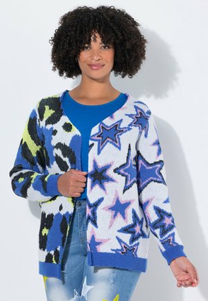 Cardigan multicolore con tonalità di blu, nero e bianco e design a stelle, con texture lavorata a maglia e polsini a costine. Indossato sopra una maglietta blu.