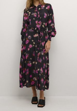 Kobieta w długiej czarnej sukience w abstrakcyjne różowe, fioletowe i beżowe plamy, z bufkami na rękawach, zapięciem na guziki z przodu i paskiem wiązanym w talii.
