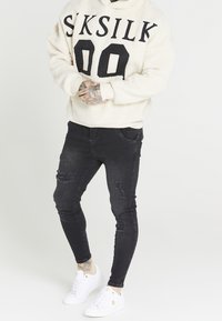 Cremefarbener Fleece-Hoodie mit schwarzem "SKSILK 00"-Aufdruck, kombiniert mit schwarzen, verwaschenen Skinny-Jeans und weißen Sneakers.