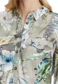 Blommig skjorta med knäppning i ett genomskinligt, lätt tyg. Har en grågrön bakgrund med mönster av blå och vita blommor samt en bröstficka.