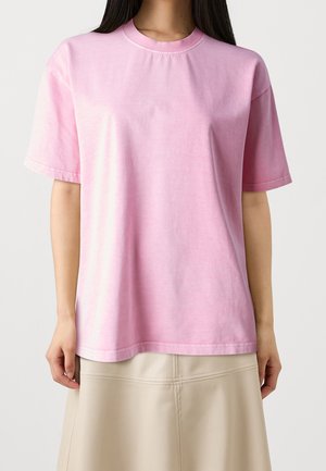 Femme portant un t-shirt rose clair uni à manches courtes et une jupe beige, debout devant un fond blanc uni.