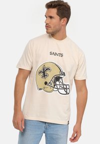 Camiseta de manga corta beige con un gráfico de un casco con una flor de lis negra y el texto "SAINTS" arriba. Fabricada en algodón, con un corte relajado.
