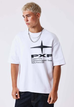 Jonge man met kort blond haar, gekleed in een wit oversized T-shirt met een zwart sterrenlogo en tekst, gecombineerd met donkere spijkerbroek en zilveren kettingen.