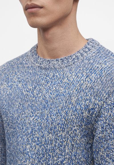 Jeune personne portant un pull tricoté bleu et blanc avec un col rond côtelé, montré du cou jusqu'au milieu de la poitrine.