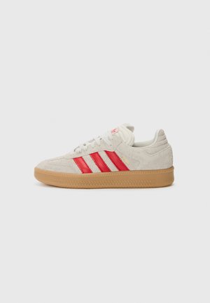 Beżowy zamszowy sneaker niskoprofilowy z czerwonymi paskami, białymi sznurowadłami i gumową podeszwą, pokazany z boku na jednolitym tle.