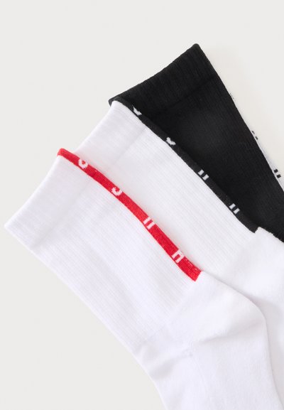 Trois paires de chaussettes montantes côtelées blanches avec bande rouge, blanches unies et noires unies, disposées en chevauchement sur un fond uni.