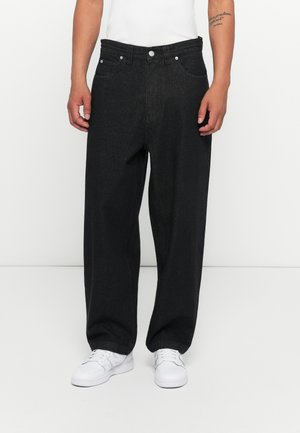 Schwarz, weit geschnittene Jeans aus Denim mit hoher Taille, Fronttaschen und Metallbeschlägen. Kombiniert mit weißen Sneakers und einem taillierten weißen T-Shirt.